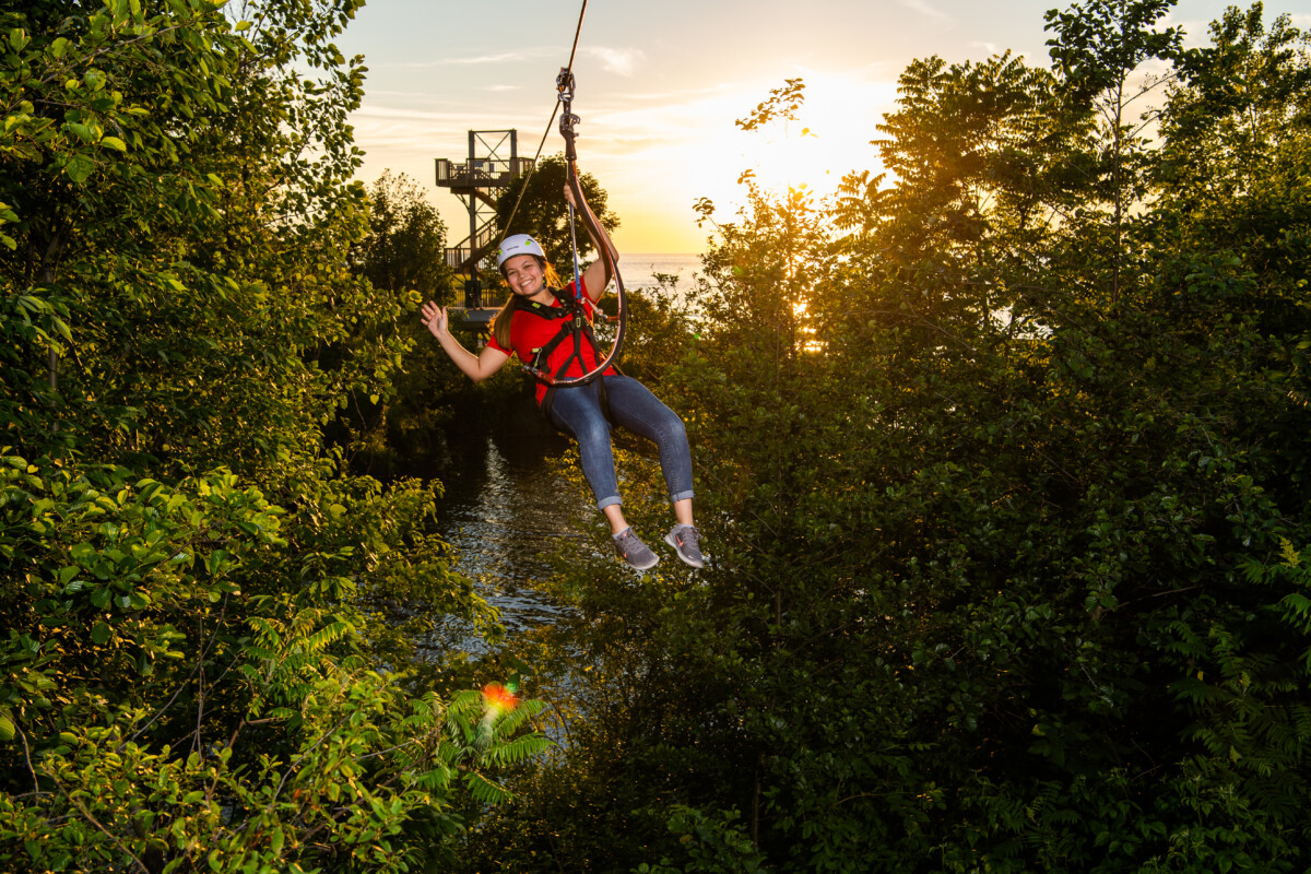 Girl zip-lining by the lake while waving and smiling.